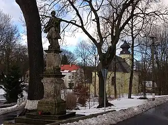 Socha sv. Jana Nepomuckého a v pozadí kostel Nanebevzetí Panny Marie v Krásném Lese