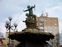 Bližší pohled na sousoší tří puttů včetně bronzové mušle zezadu. Stojící amorret má malá křídla. V pozadí za sousoším jsou vidět tři panelové domy