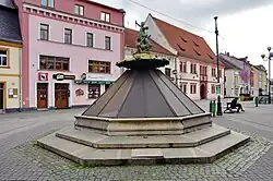 Pohled na kašnu, která je opatřena dřevěným bedněním. Vidět jde pouze sousoší puttů. Za ní jsou vidět domky na Starém náměstí, včetně původní radnice