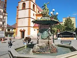 Bližší pohled na sloup kašny a sousoší puttů. Na sloupu lze vidět bronzový reliéf Augusta Schrama. V pozadí stojí kostel sv. Jakuba Většího