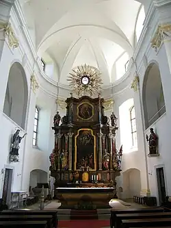 Barevná fotografie, uprostřed hlavní oltář s titulárním obrazem a sochami, po stranách sochy, v popředí lavice