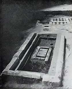 Černobílá fotografie. V obdélníkovém hrobu leží schránka.