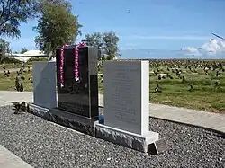 Barevná fotografie památníku na pobřeží ostrova. Památník má podobu tří pamětních desek vedle sebe na nízkém soklu. Prostřední je z černé leštěné žuly, krajní z bílého kamene. Na deskách je výčet amerických jednotek účastnících se bitvy a mapka bitvy.