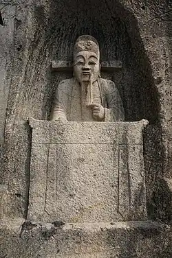 fotografie kamenné sochy vytesané do skalního výklenku, vytesán je muž sedící za stolem, v úředním rouchu a klobouku, levou rukou si hladící vous