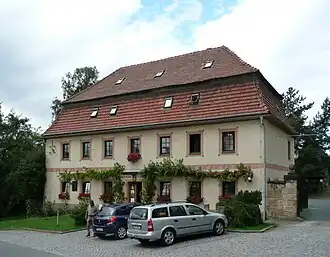 Světlehnědá budova s červenou mansardovou střechou, v popředí dláždění, dvě auta a vedle stojící osoba.