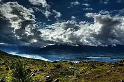 Pohled na tmavě modré jezero, vepředu sluncem nasvícená vegetace, vzadu mraky na temné obloze