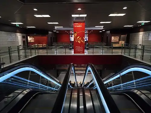 Vestibul podzemní stanice metra. Uprostřed v popředí dvojice eskalátorů, které vedou dolů na nástupiště. Strop z panelů s integrovanými světly. V pozadí na zdech panely s informacemi o archeologických nálezech s fotografiemi. Interiér je laděn v šedé a červené barvě.