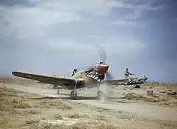 Kittyhawk Mk.III 112. peruti při pojíždění v Medeníně v Tunisku, 1943. Příslušník pozemního personálu sedící na křídle navádí pilota jehož výhled vpřed je omezen předkem letadla.