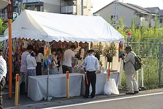 Pohled na provizorní bílý stánek, postavený vedle silnice, se spousty květin a malého oltáře, kde muži a ženy kloní své hlavy k uctění památky obětí