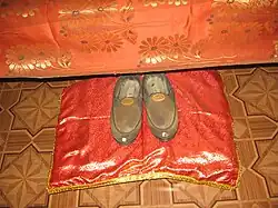 Fotografie: střevíce duchovního učitele (guru) jako předmět úcty uložené na zdobný polštářek u postele v místnosti jíž obýval.