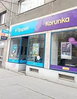 Pohled na prodejnu v přízemí vícepatrové budovy. Nad pobočkou je modrá cedule s nápisy Tipsport a Korunka, na výloze jsou reklamy na produkty společnosti, v popředí je větev stromu bez listí.