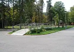 Pohled do parku. V parku jsou vedeny dvě asfaltové cesty, které se vpředu spojují. Mezi cestami se nachází travnatý pás. Na tomto pásu, rovnoběžně s oběma cestami, se klene nízký most, na jehož zábradlí jsou tabulky se jmény dekorovaných osobností.