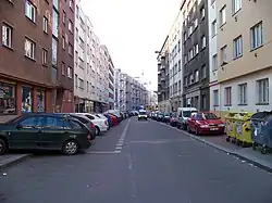 úzká ulice s pětipatrovými obytnými domy po obou stranách, před domy zaparkované automobily