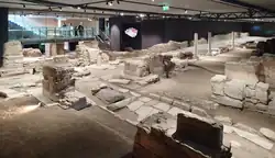 Prostory vestibulu stanice metra. Na obrázku jsou vidět odhalené antické archeologické památky, včetně kamenných zdí, sloupů a dlažeb. Vše je zasazené do moderní betonové konstrukce stanice. V pozadí je vidět, že nad archeologickým nalezištěm visí kovová plošina, schodiště a lávka pro návštěvníky. Na stropě visí řada světel, které prostor osvětlují.