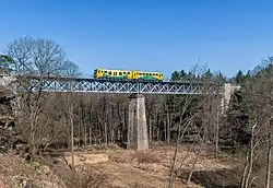 Viadukt přes údolí Peklo nedaleko Zahrádek u České Lípy, postavený kombinací kamenných oblouků a ocelové příhradoviny. Na předjarním záběru vystupují obnažené skalní masivy, kterými trať prochází, a stromy postrádající olistění. Po mostě projíždí motorová jednotka řady 814, takzvaná Regionova.