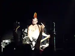 Vladimír Šťástka s drsným výrazem a bez trička sleduje své publikum během hraní; na hlavě má tradiční žlutočervené papírové číro a v popředí v šeru je pogující dav