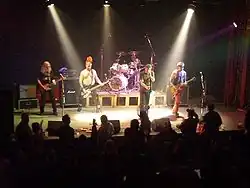 Záběr na osvětlené pódium s kapelou Visací zámek, Haubert uprostřed právě zpívá, Pixa je při pohledu diváka vpravo od něj, Pátek je za ním za bicími, vlevo pak Šťástka a na kraji pódia Rut; v popředí pulzuje publikum