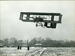 Fotografie dvojplošníku Voisin ve vzduchu. Pilotem je pravděpodobně Robert Svendsen, i když to nelze s jistotou potvrdit. 1910.