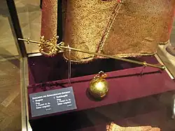 Barevná fotografie pozlaceného královského žezla a jablka vystavených ve vitríně ve vídeňské klenotnici. V pozadí je vidět spodní část kurfiřtského pláště.