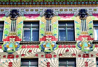 Fasáda budovy ve stylu vídeňské secese (Jugendstil) s bohatě zdobeným průčelím v jasných barvách. Na fasádě jsou tři okna s charakteristickými barevnými květinovými a geometrickými motivy v červené, zelené, modré a žluté barvě.