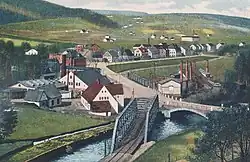 Pohled na německý Grünthal. Flájská dráha (Flöhatalbahn) přechází Flájský potok. Zaniklá obec je mimo pohlednici vpravo