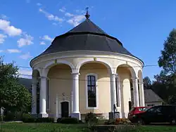 Kruhová stavba kostela. Budova je ve světle žluté barvě a její střecha je tmavá. Před kostelem je zelený trávník, na němž na snímku vpravo parkují osobní automobily.