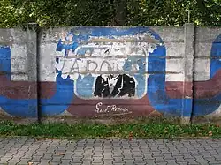 Betonová zeď, za ní vykukují ze zahrady zelené stromy. Před zdí vede chodník. Na zdi je namalovaný, leč vandaly poničený, obraz Ľudevíta Vladimíra Riznera.