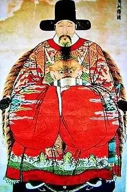 Postarší muž sedící v křesle, oblečený v honosném hávu s typickými čínskými širokými rukávy, na hlavě má úřednickou čapku.